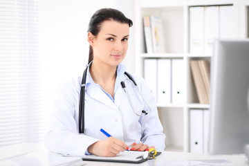 Young brunette female doctor sitting  with clipboard near window in  hospital and filling up medical history form.