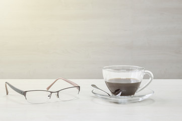 Closeup black coffee in transparent cup of coffee and eyeglasses on blurred wooden desk and wall textured background , relaxation in coffee break concept by coffee and eyeglasses in art tone