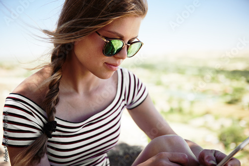 Fototapeta na szkło Zakończenie piękna młoda kobieta w eleganckich okularach przeciwsłonecznych texting wiadomości na jej mądrze telefonie. Ładna kobieta networking outdoors w letnim dniu.