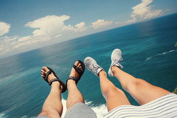 Legs of man in sandals and woman in sport shoes sitting above the ocean