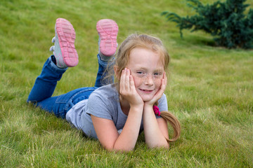 little ginger girl with freckles is laying on the lawn, head is leaning on the hands