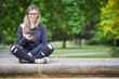 © nenadaksic - Young woman sitting on the wall in the park, writing a message on her mobile phone, park in the background