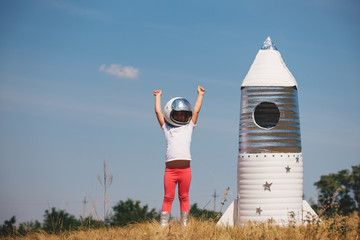 Happy child girl dressed in an astronaut costume playing with hand made rocket. Summer outdoor