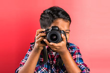 10 Year Old Indian Boy Holding Digital Camera Or DSLR Camera, Posing Like A Professional Photographer, Young Photographer, Kid Photographer, Child Photographer, Portrait, Closeup, Red Background