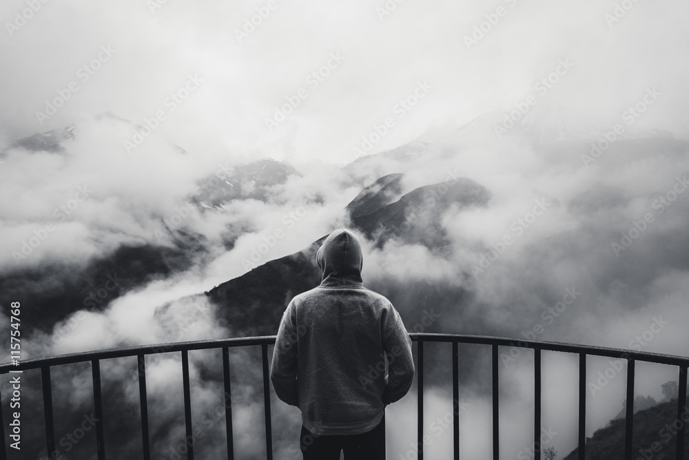 View from behind of a man standing at view point looking to beautiful landscape with foggy mountains in the distance. Black and white photo.