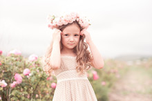 Summer Portrait Of Baby Girl Wearing Trendy Dress And Hairstyle With Roses Outdoors. Looking At Camera.