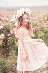 Canvas Print - Smiling kid girl 4-5 year old posing in rose garden outdoors. Wearing stylish dress and wreath with flowers. Summer portrait