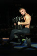 © kazanovskyiphoto - Handsome muscular man sitting with dumbbells on dark  background