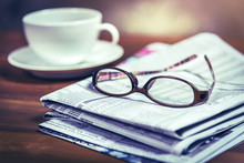 Glasses On Pile Of Newspaper With Coffee Cup In Background.Vintage Tone Photo With Selective Focus On Glasses.
