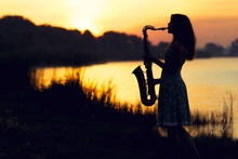 Silhouette Portrait Of A Woman In A Dress Whose Hobby Music, She Plays On The River Bank And Is Resting Emotionally