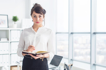 Wall Mural - Beautiful businesswoman standing in office, holding notebook, planning meetings for the work day, looking at camera.