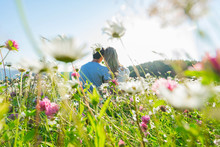 Couple Sitting In The Flowers Field. Selective Focus: On The Few Flowers In The Middle. Flowers On The Front Of Picture And Couple Are Blured.