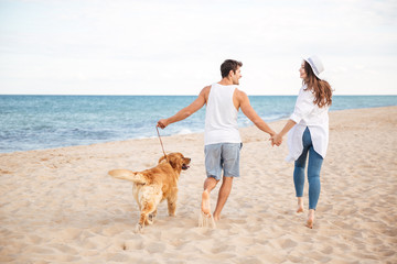 Canvas Print - Happy joyful young couple running on beach with their dog