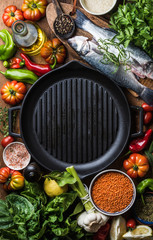 Wall Mural - Raw uncooked seabass fish, vegetables, grains, herbs, spices and olive oil on chopping board, iron grilling pan in center with copy space, top view