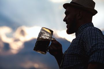 Happy smiling man tasting fresh brewed beer against the sky at sunset. The theme is Oktoberfest, a guy in Bavarian style