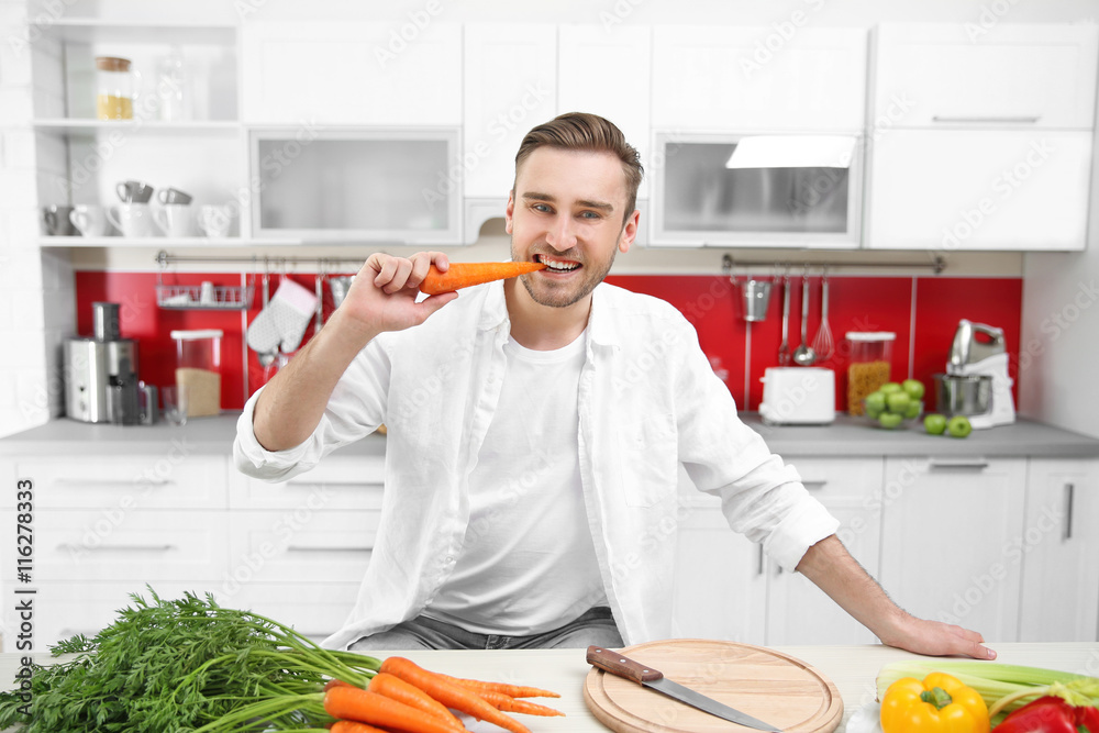 Handsome man eating carrot in kitchen