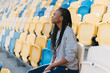 Excited african american woman in gray sportswear looking away sitting on bleachers waiting for a show, celebration
