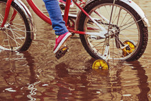 Child On Bike Rides Through A Puddle In Autumn Day.