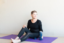 Older Woman In Black Yoga Clothing Smiling And Relaxing On Purple Mat With Blue Glass Water Bottle After Yoga
