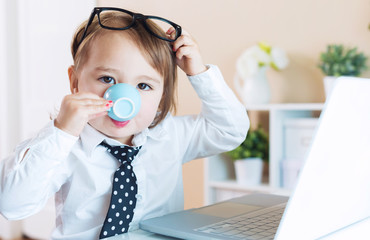 Wall Mural - Smart toddler girl with glasses drinking coffee while using a laptop