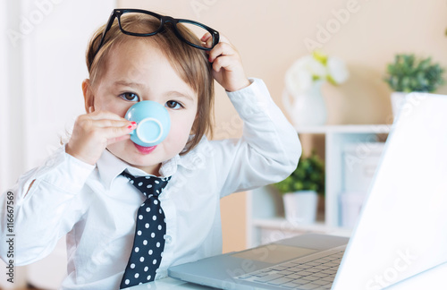 Photo Smart toddler girl with glasses drinking coffee while using a laptop