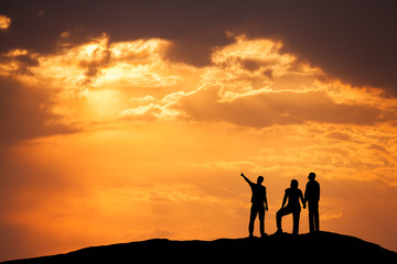 Landscape with silhouette of a standing man pointing finger in colorful sky and his friends on the high mountain peak against the beautiful sunset. High rocks. Travel, Climbing, Trekking.