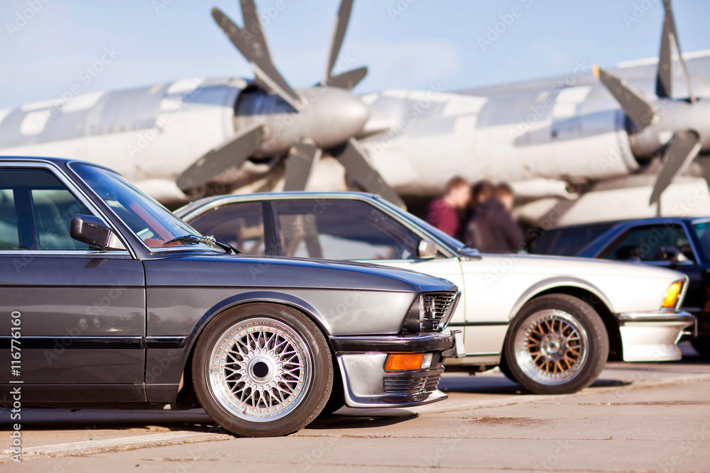 Right front side of two old european sport cars with plane on background - obrazy, fototapety, plakaty 