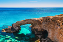 Woman On The Beautiful Natural Rock Arch Near Of Ayia Napa, Cavo Greco And Protaras On Cyprus Island, Mediterranean Sea. Legendary Bridge Lovers. Amazing Blue Green Sea And Sunny Day.