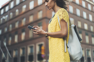 Young hipster girl wearing yellow dress and using modern smartphone outside, attractive smiling woman typing text message on her cellphone outdoors, social networking concept