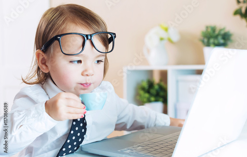 Photo Smart toddler girl with glasses drinking coffee while using a laptop