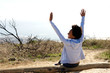 © mimagephotos - Young woman sitting on log with her hands raised