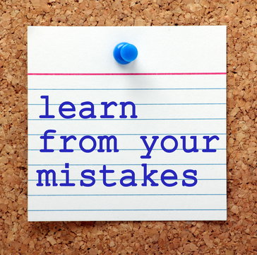 The words Learn From Your Mistakes in blue text on a note card pinned to a cork notice board as a reminder