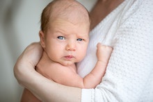 Portrait Of Cute Newborn Baby Looking At Camera With Funny Serious Expression While Sitting On Mothers Arms. Young Mom Holding Healthy New Born Child. Love, Childcare, Happy Family Concepts