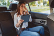 © perfectlab - Young, beautiful woman sitting in the back seat of the car with a tablet in hand and drinking coffee