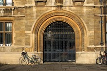 Parked Bikes Leaning Against Historic Stone Building Of Gonville And Caius College, Cambridge, By Ornamental Iron Gate.