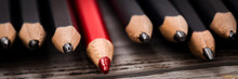 Red Pencil Stands Out From The Crowd Of Black  On A Wooden White Background.