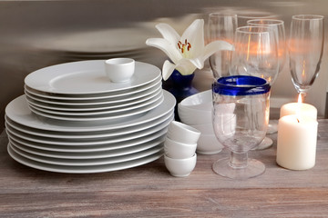 Wall Mural - White dishes stacked on a wooden table against steel background with transparent wineglasse. flower and burning candles. 