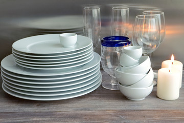 Wall Mural - White dishes stacked on a wooden table against steel background with transparent wineglasses. Toned image. Composition with burning candles.