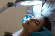Procedure of facial skin examination at cosmetologist's. Portrait of a young woman with closed eyes and specialist's hands in medical gloves under magnifying lamp. Close up. Indoor shot.