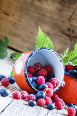 Raspberries and blueberries in red enamel mug, old wooden backgr