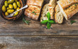 Green mediterranean olives in bowl and slices of freshly baked ciabatta over rustic wooden background. Top view, copy space, horizontal composition