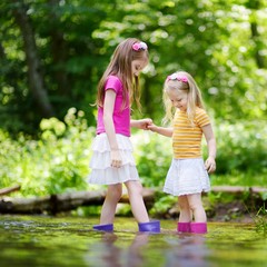 Wall Mural - Two cute little sisters playing in a river wearing raining boots