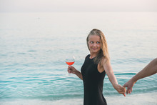 Young Blond Woman With Glass Of Rose Wine Holding Man's Hand On Beach By The Sea At Sunset. Kleopatra Beach, Alanya, Mediterranean Region, Turkey.
