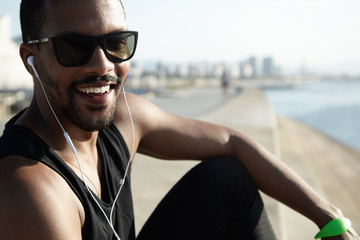 Portrait of good-looking young African athlete wearing stylish shades and black outfit, looking happy and cheerful, sitting on sidewalks at sea, waiting for his friend to join him in evening run