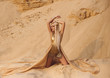 Beautiful queen of the desert sitting on the sand. Luxury golden dress and unusual jewelry. Gold flower in her hair, hairstyle with braids. Beautiful blonde. Fashionable toning.