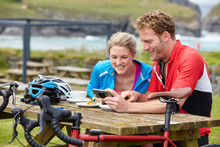 Cyclists Using Mobile Phone At Picnic Table Overlooking Ocean