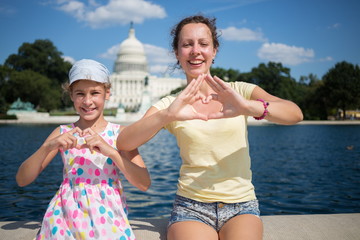 Woman with daughter depict hearts near pond and Capitol at summer.