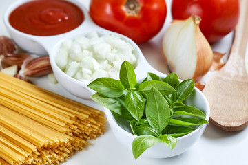 Basil, sliced onions and tomato paste in white bowl with raw spaghetti and garlic scattered around on white background