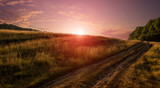 Fototapeta  - Field and a dirt road on the sunrise. magnificent picturesque scene. majestic sunset, amazing rural landscape