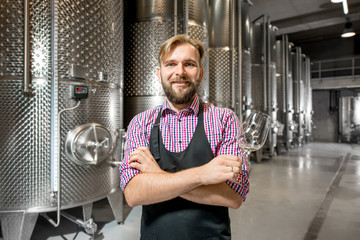 Portrait of a handsome wine maker in working apron with the wine glass at the manufacture with metal tanks for wine fermentation. Wine production at the modern factory